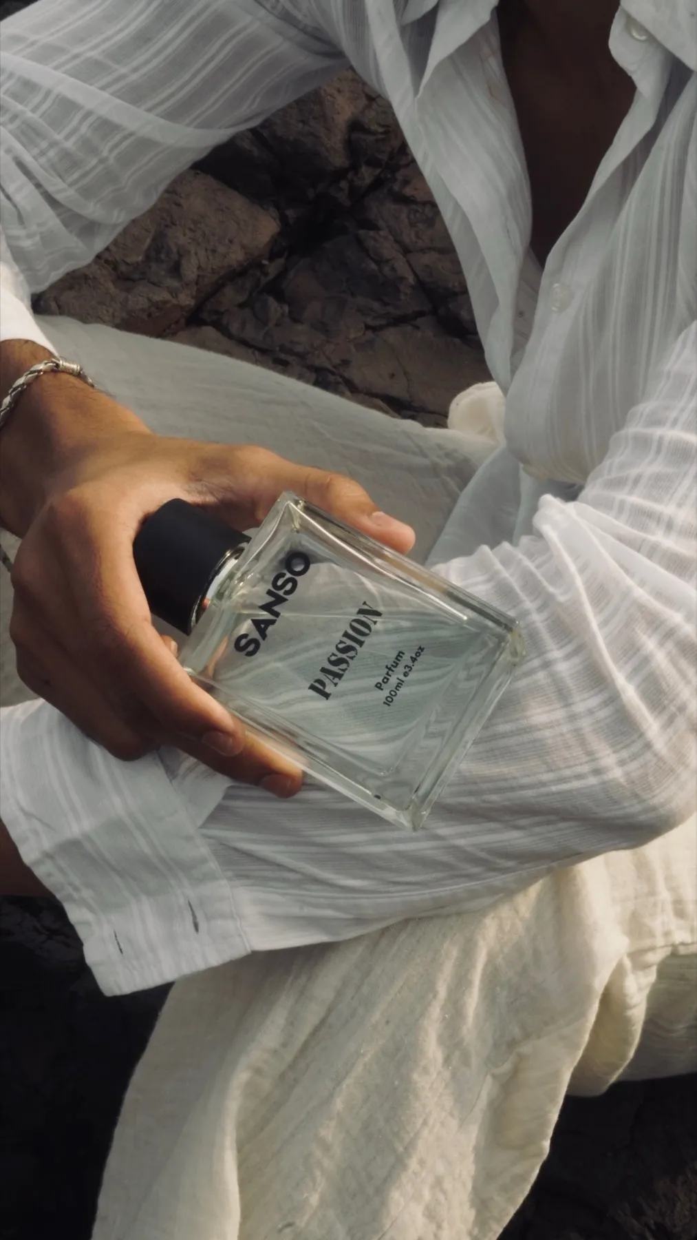 Sanso Passion parfum bottle held in one hand against a white linen shirt, on black sea rock